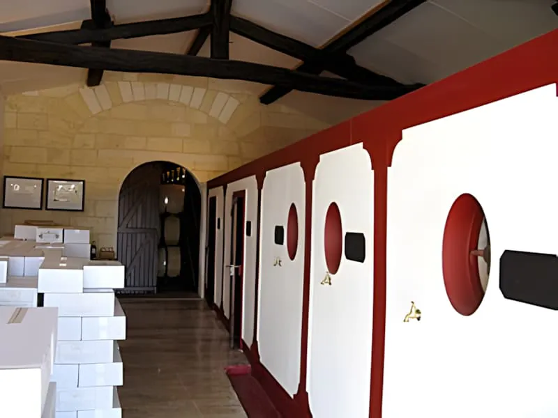 Intérieur d'une salle de vente de vin avec des murs blancs, des bordures rouges, des poutres en bois apparentes, et des certificats encadrés sur les murs avec une porte d'entrée en bois.