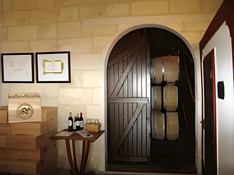 Une vue plus rapprochée de l'entrée de la salle de vente montrant la porte distinctive en bois, l'arcade en pierre, et la table d'exposition de vin avec des certificats montés sur le mur.