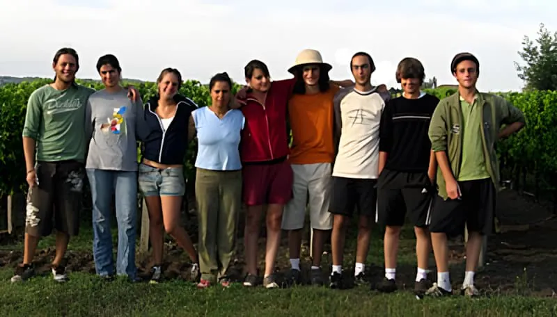 A diverse group of nine temporary workers standing together in a line in front of a vineyard at Château Plaisance, smiling for a group photo.