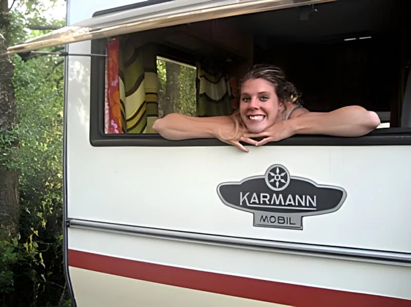 Eine lächelnde junge Frau mit blonden Haaren lehnt sich aus was wie ein Wohnmobil oder Anhängerfenster aussieht.