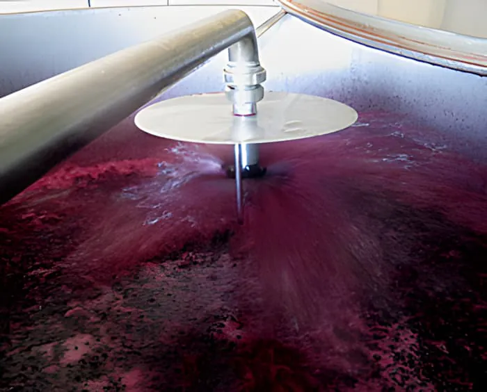 深红色葡萄酒被泵送并喷洒在大型不锈钢发酵罐中葡萄固体顶部的特写。