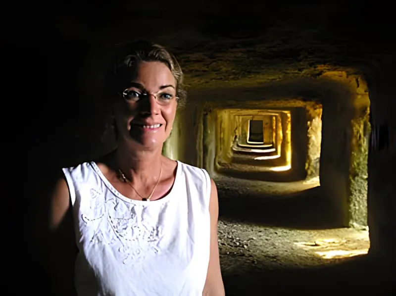 A woman in white standing in a carved limestone tunnel with warm ochre-colored walls, with additional chambers visible stretching into the distance.