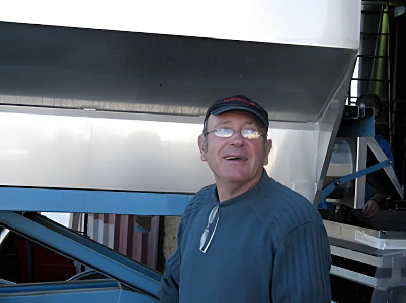 A smiling middle-aged man wearing glasses and a dark cap, photographed in what appears to be a workshop or storage area.
