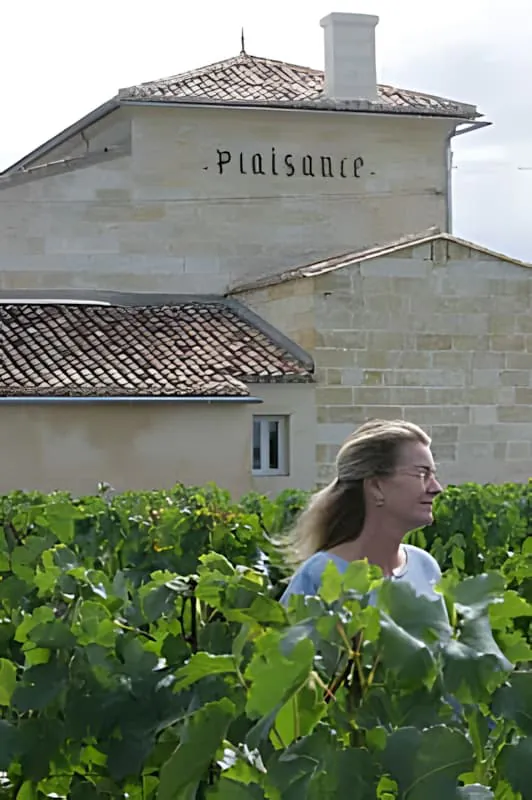 A woman with blonde hair working among lush green grapevines with the white stone Château Plaisance building visible in the background.
