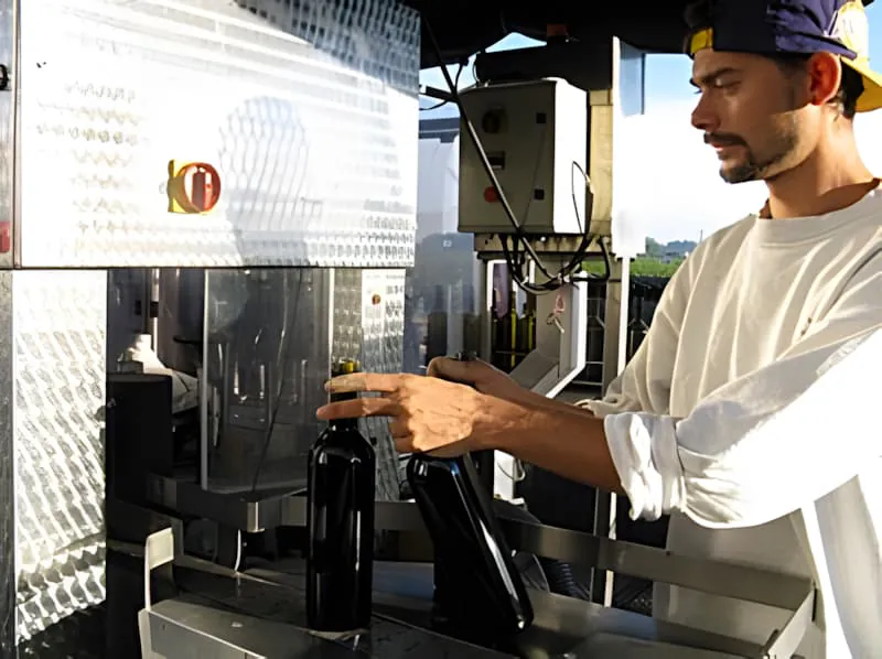 一名戴帽子的工人在装瓶线上将装满的酒瓶放入金属储存箱,显示过程的完成一边。